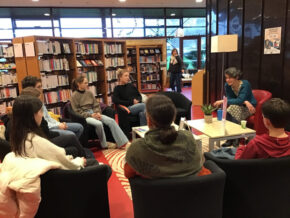 Dans une bibliothèque, une femme, assise dans un fauteuil, parle à sept personnes assises en cercle autour d'elle. Des rayonnages de livres sont situés derrière elles.