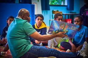 Un homme assis lit un livre à des enfants devant lui qui sont également assis.