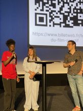 Deux adolescentes se tiennent sur une scène aux côtés du bédéiste Charlie Zanello. L'une d'entre elles semble parler dans un micro.