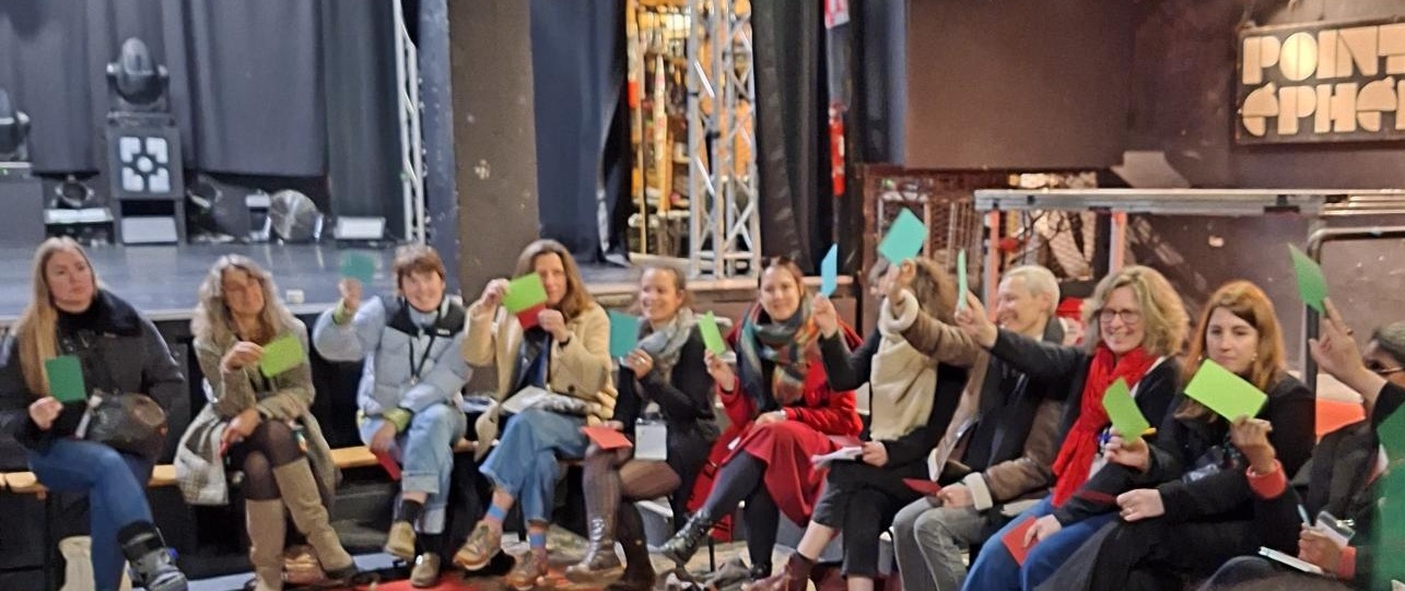 Une dizaine de femmes sont assises sur des chaises disposées en demi-cercle dans une salle de spectacle. Souriantes, elles brandissent des post-it. 
